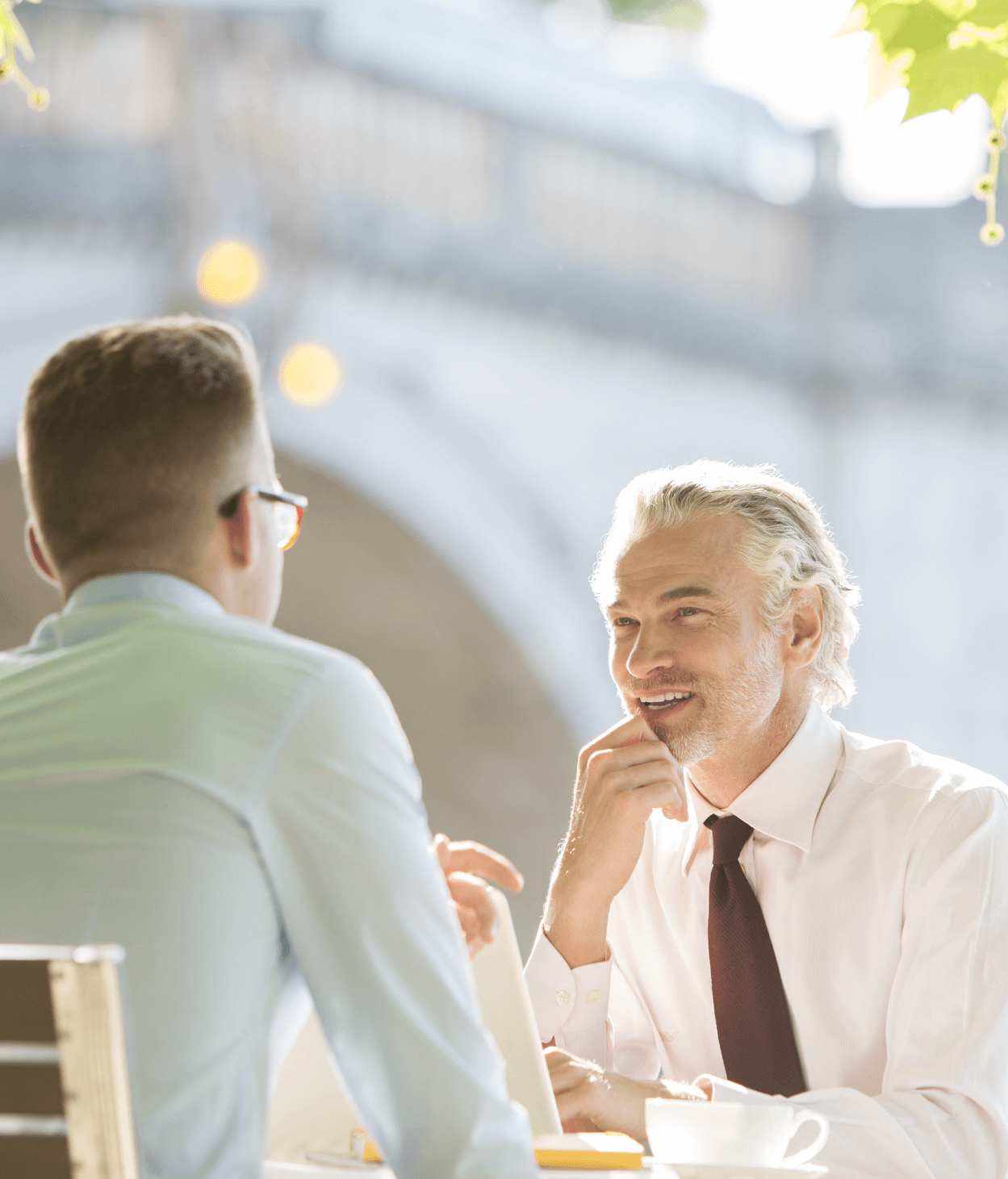 photo of two men dressed in business attire chatting at a table outside of a cafe.