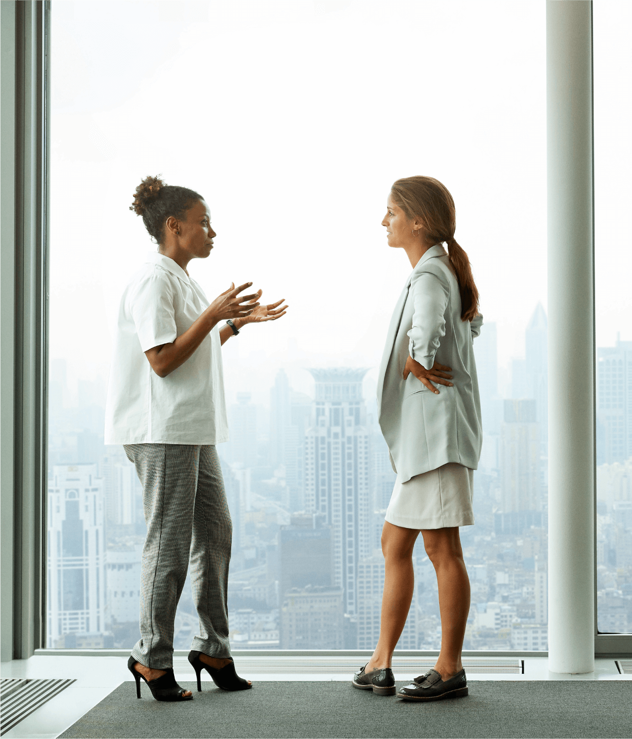 Photo of two female coworkers speaking by a window overlooking a city